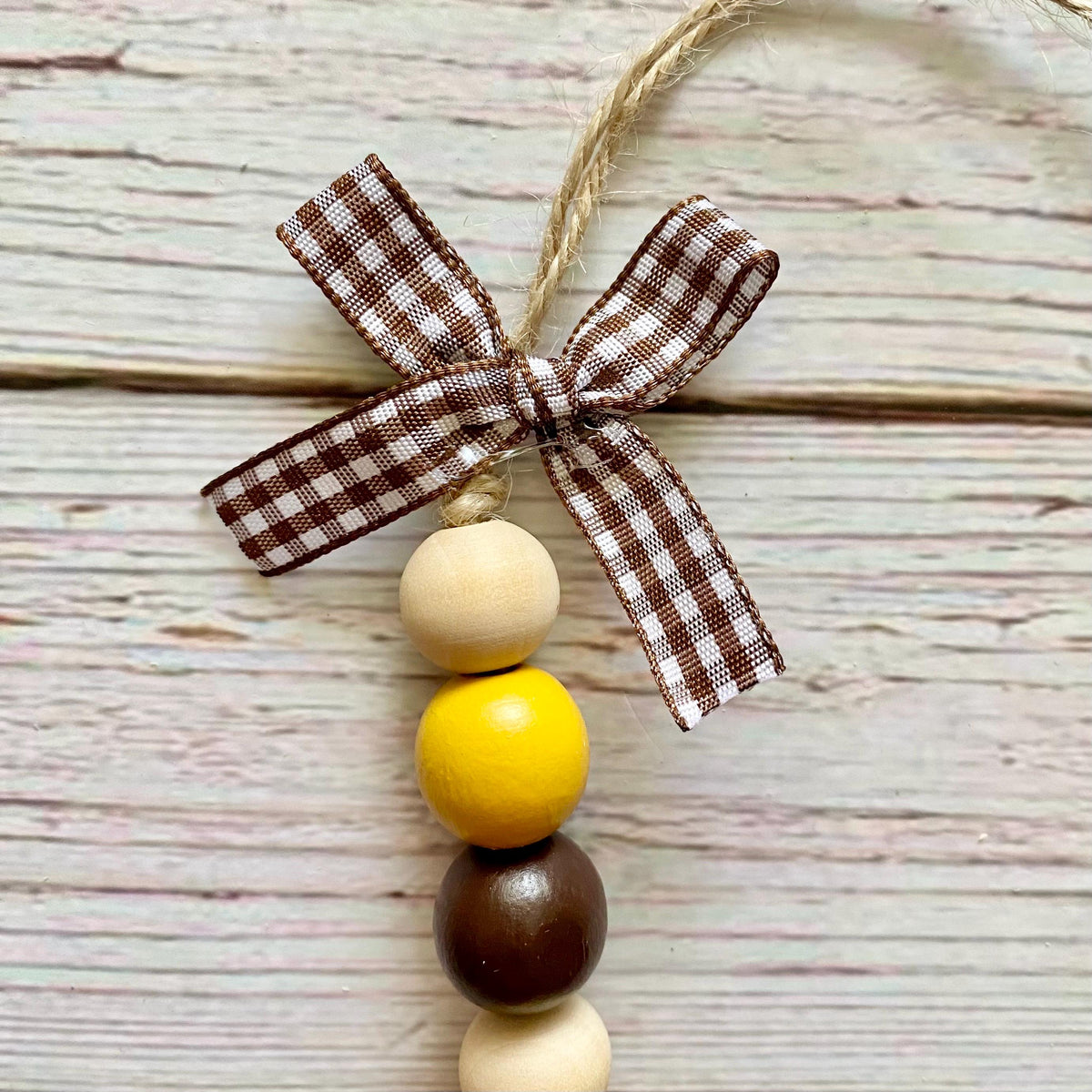 A rustic wooden bead garland ornament featuring a brown and white gingham ribbon bow at the top, hanging from a jute string. The ornament has four wooden beads: two natural cream, one bright yellow, and one dark brown, arranged vertically on a whitewashed wood plank background.