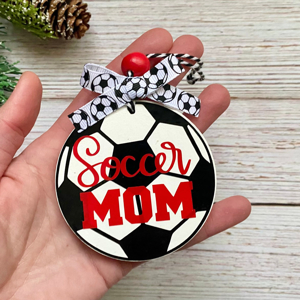 A hand holds a round soccer ornament featuring a black and white soccer ball design with the words 'Soccer MOM' in red script and block letters. The ornament has a black, white, and gray patterned ribbon loop with a small red bead at the top, set against a light wood background with pine branches in the corner.