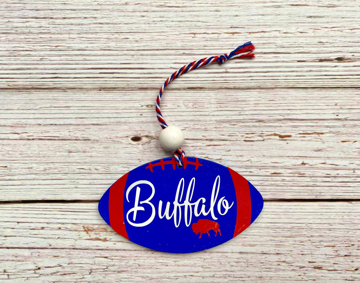 A football-shaped wooden ornament with a blue and red Buffalo Bills design, featuring the word 'Buffalo' in white script and a small red buffalo silhouette. It hangs from a red, white, and blue braided cord with a white bead, displayed on a rustic white-washed wooden background.