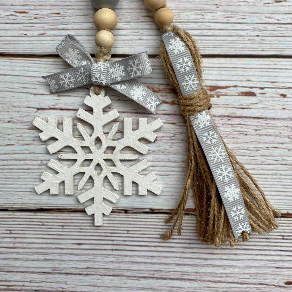 A decorative hanging ornament set on a whitewashed wood plank background, featuring a white snowflake charm, a gray ribbon bow with snowflake pattern, natural wood beads, and a matching tassel with jute rope and gray ribbon.