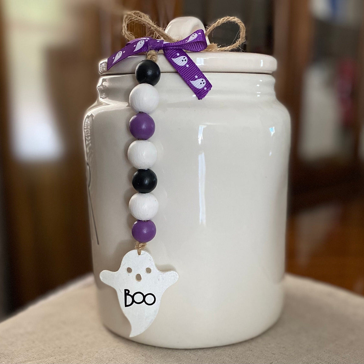 A white ceramic jar with a matching lid, decorated with a purple ribbon bow, a beaded garland in black, white, and purple, and a white ghost charm that says 'Boo' in black lettering. The jar is sitting on a neutral fabric surface with a blurred indoor background.