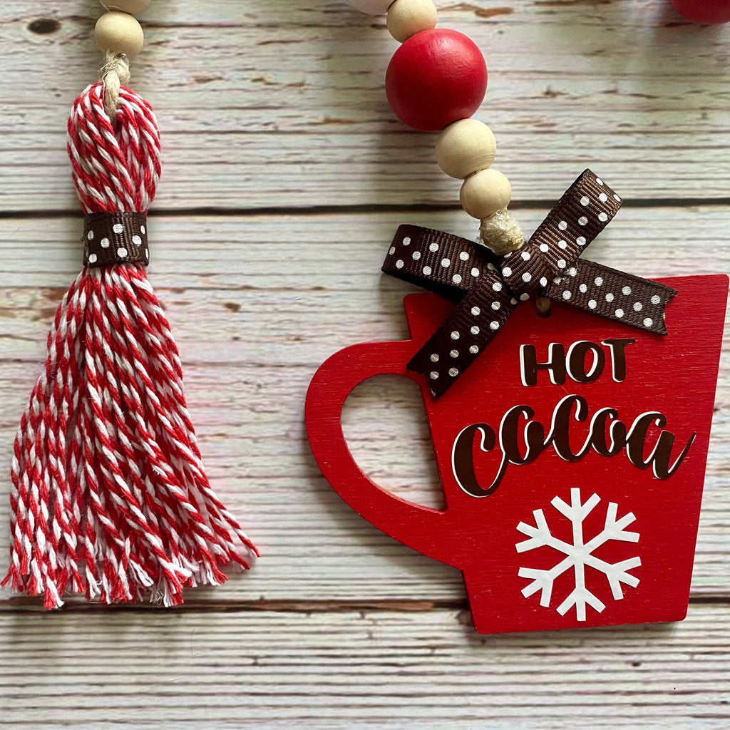 A close-up of a holiday hanging decoration featuring a red wooden mug with 'HOT COCOA' and a snowflake, tied with a brown polka-dot ribbon, next to a red and white striped tassel bead garland on a whitewashed wood background.