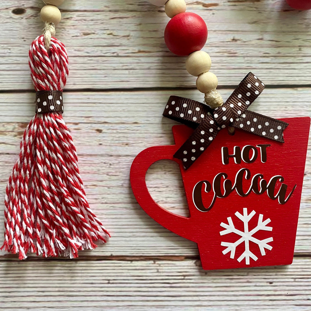 A close-up of a holiday hanging decoration featuring a red wooden mug with 'HOT COCOA' and a snowflake, tied with a brown polka-dot ribbon, next to a red and white striped tassel bead garland on a whitewashed wood background.