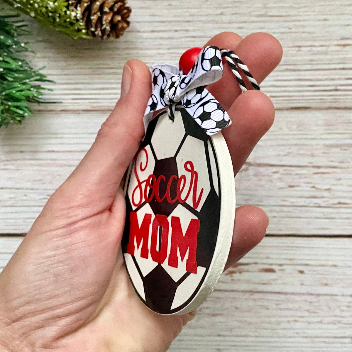 A hand holds a decorative oval ornament shaped like a soccer ball, with the words 'Soccer MOM' in red and black. The ornament has a black-and-white patterned ribbon loop with a small red bead, shown against a light wood plank background with a pine branch and pinecone as props.