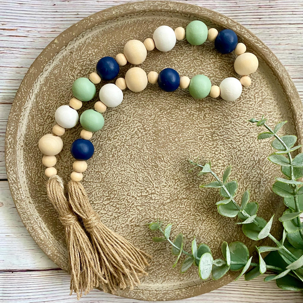 A beaded garland with natural wood beads, painted light green, white, and navy blue beads, and two tan jute tassels, arranged on a round textured beige tray with eucalyptus leaves.