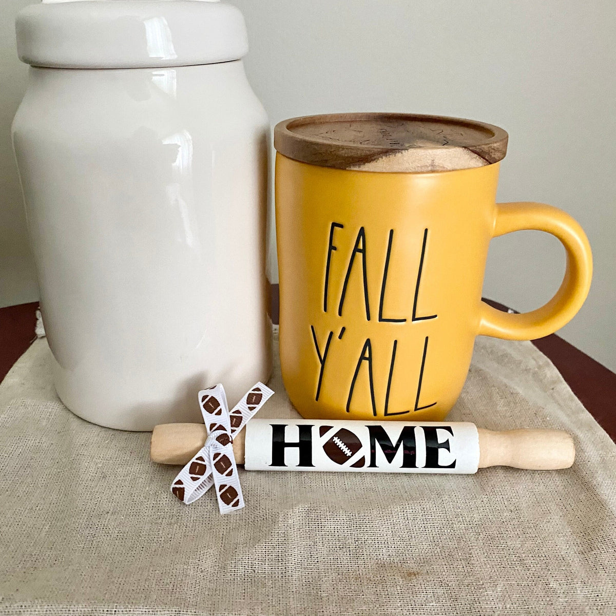 A yellow ceramic mug with a wooden lid and the words 'FALL Y'ALL' in black lettering, styled on a beige cloth next to a white jar and a wooden rolling pin with 'HOME' written on it.