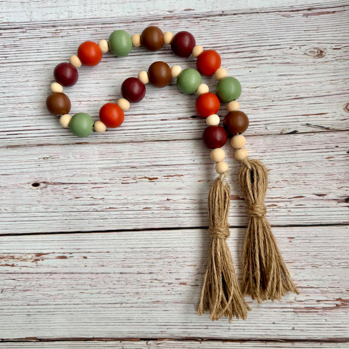 A beaded garland with alternating painted wooden beads in red, green, brown, and cream, finished with two knotted jute tassels, displayed on a rustic white-washed wood background.