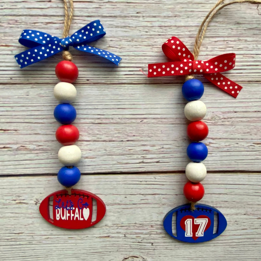 Two patriotic football-themed wooden bead ornaments on a whitewashed wood background. One ornament is red with a blue polka-dot bow and a red football-shaped tag reading 'Buffalo'.