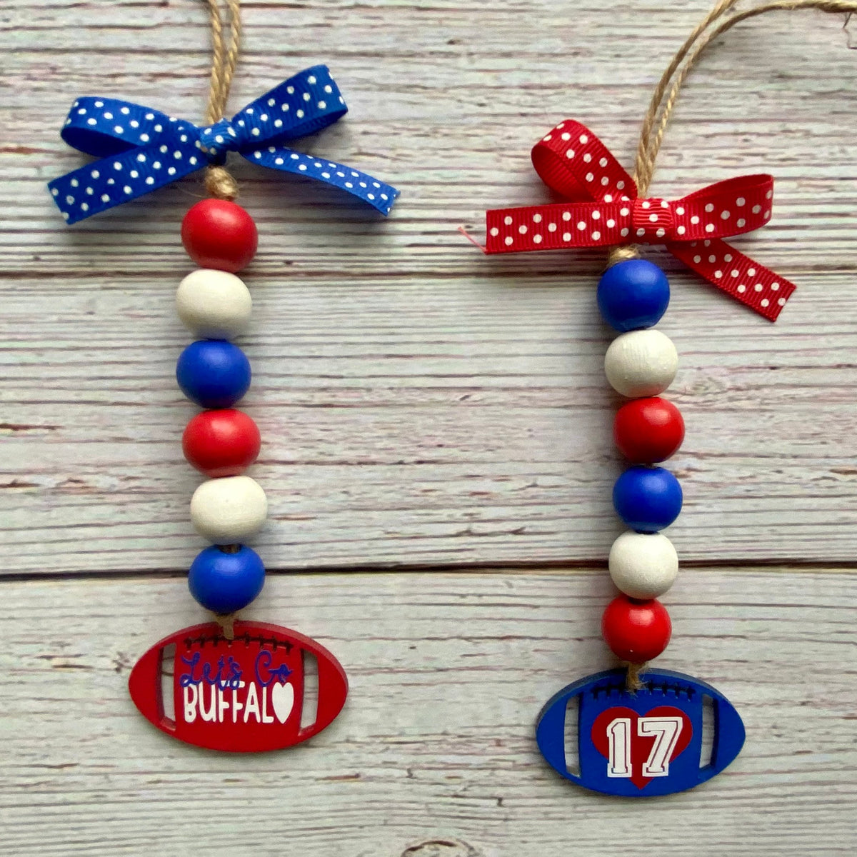 Two patriotic football-themed wooden bead ornaments on a whitewashed wood background. One ornament is red with a blue polka-dot bow and a red football-shaped tag reading 'Buffalo'.