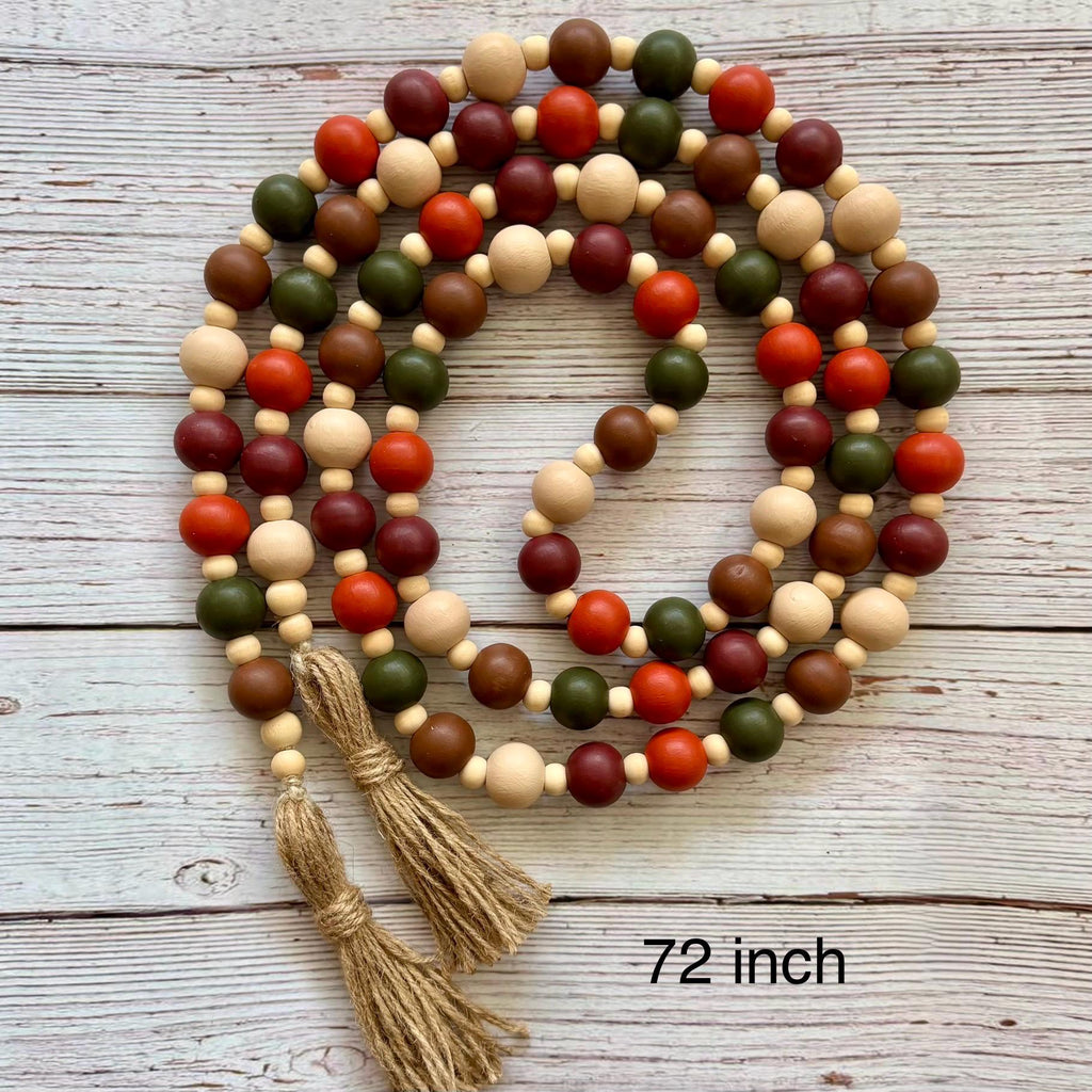 A wooden bead garland with alternating natural beige, dark red, orange, and olive green beads, finished with two knotted jute tassels, displayed on a light gray wood plank background with a '72 inch' text overlay.