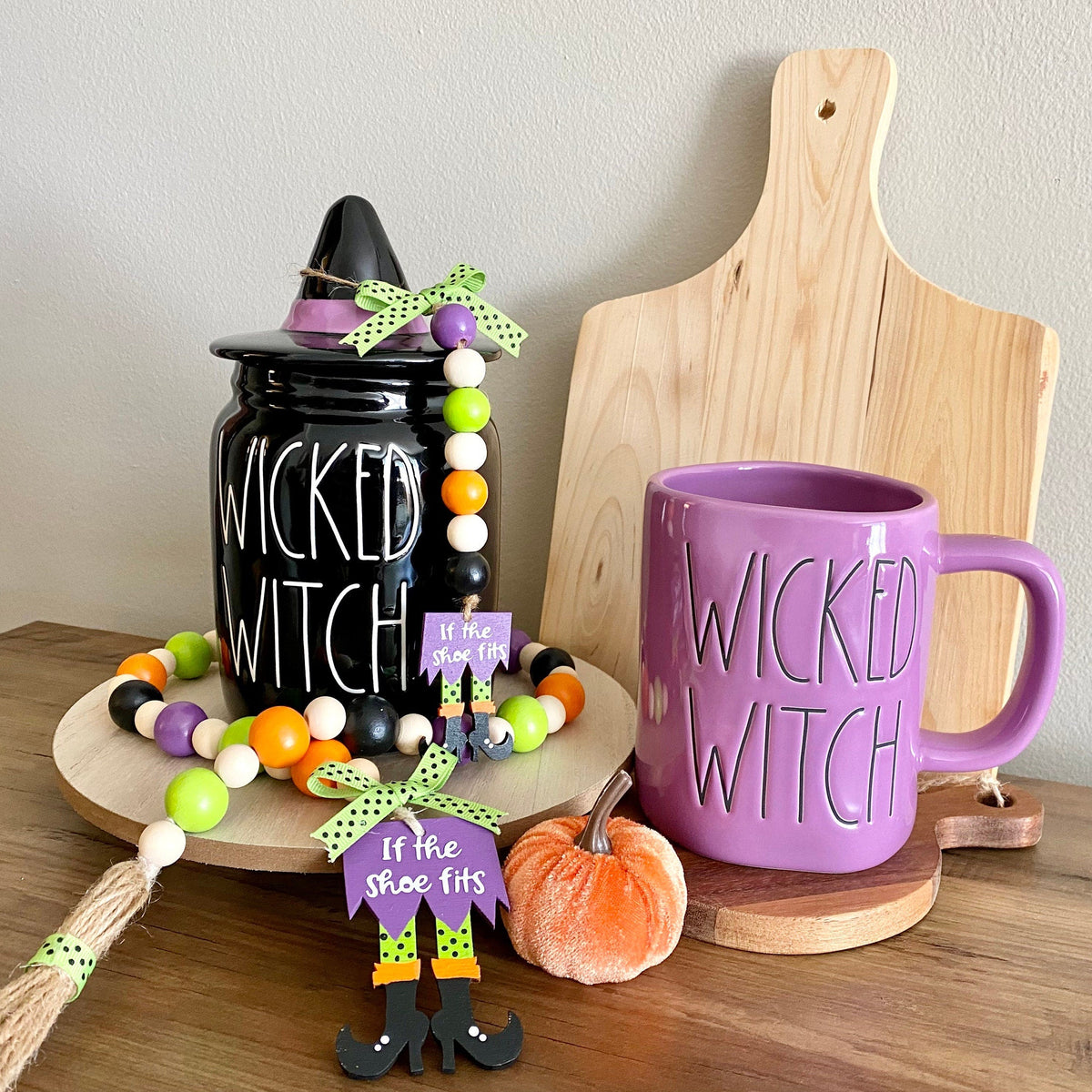 Wicked Witch Halloween-themed tabletop decor set featuring a black ceramic jar with a witch hat lid, a purple ceramic mug, wooden beads, a small orange pumpkin, and a wooden cutting board on a wooden table.