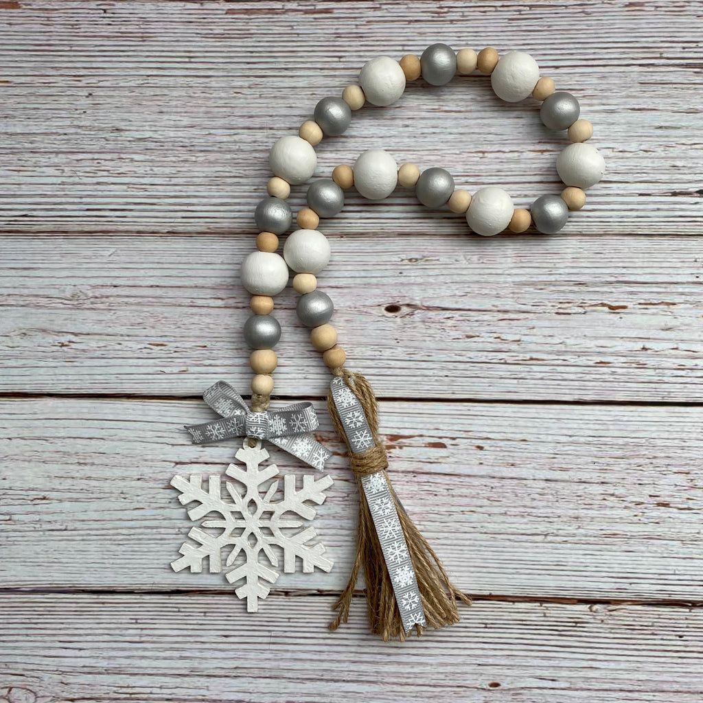 A wooden bead garland with white, gray, and natural wood beads, featuring a white snowflake ornament, a silver and white patterned ribbon bow, and a matching tassel, displayed on a rustic white-washed wooden background.