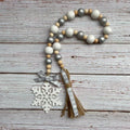 A wooden bead garland with white, gray, and natural wood beads, featuring a white snowflake ornament, a silver and white patterned ribbon bow, and a matching tassel, displayed on a rustic white-washed wooden background.