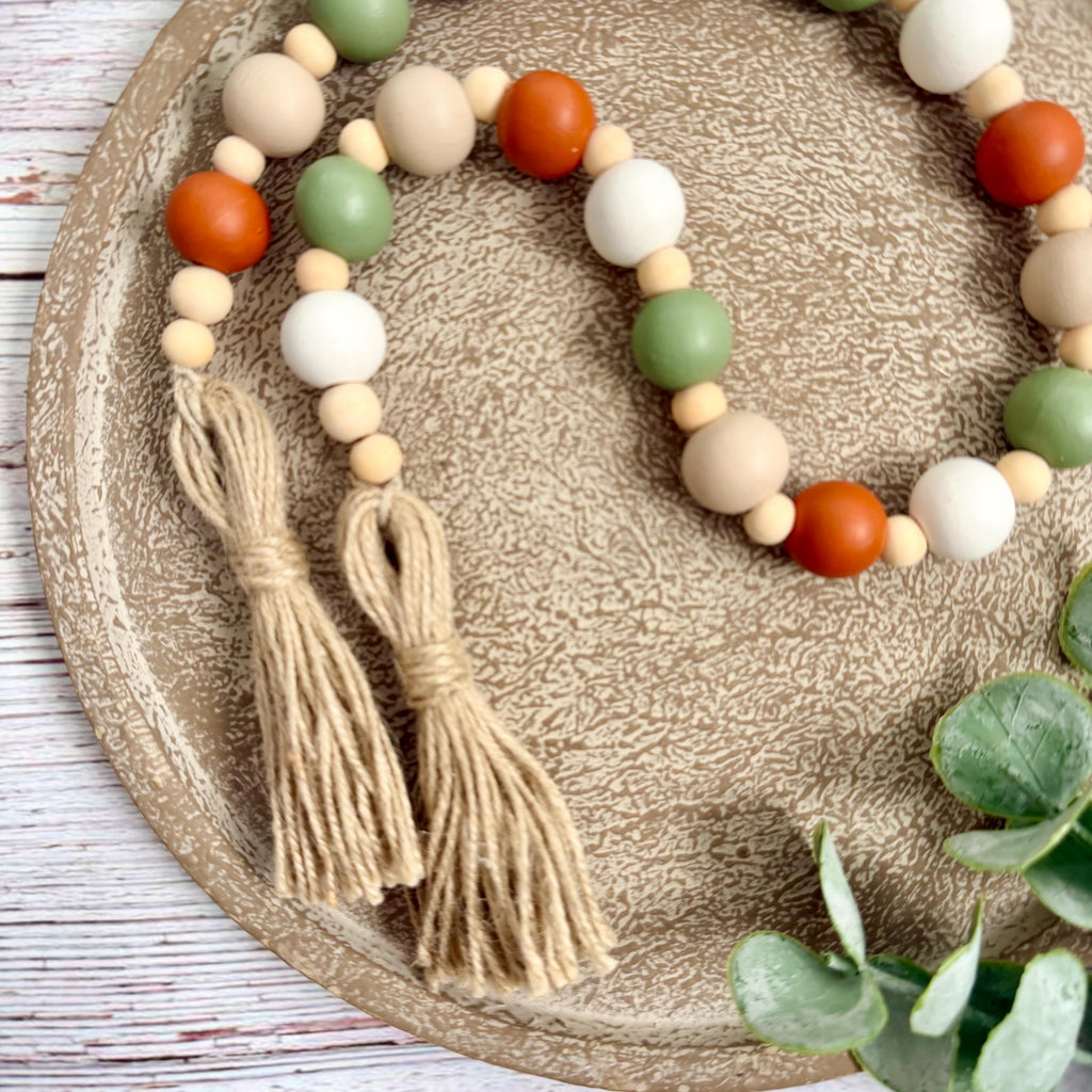 A beaded garland with two tassels, featuring matte wooden beads in terracotta orange, sage green, cream, and white, displayed on a textured beige tray with eucalyptus leaves.