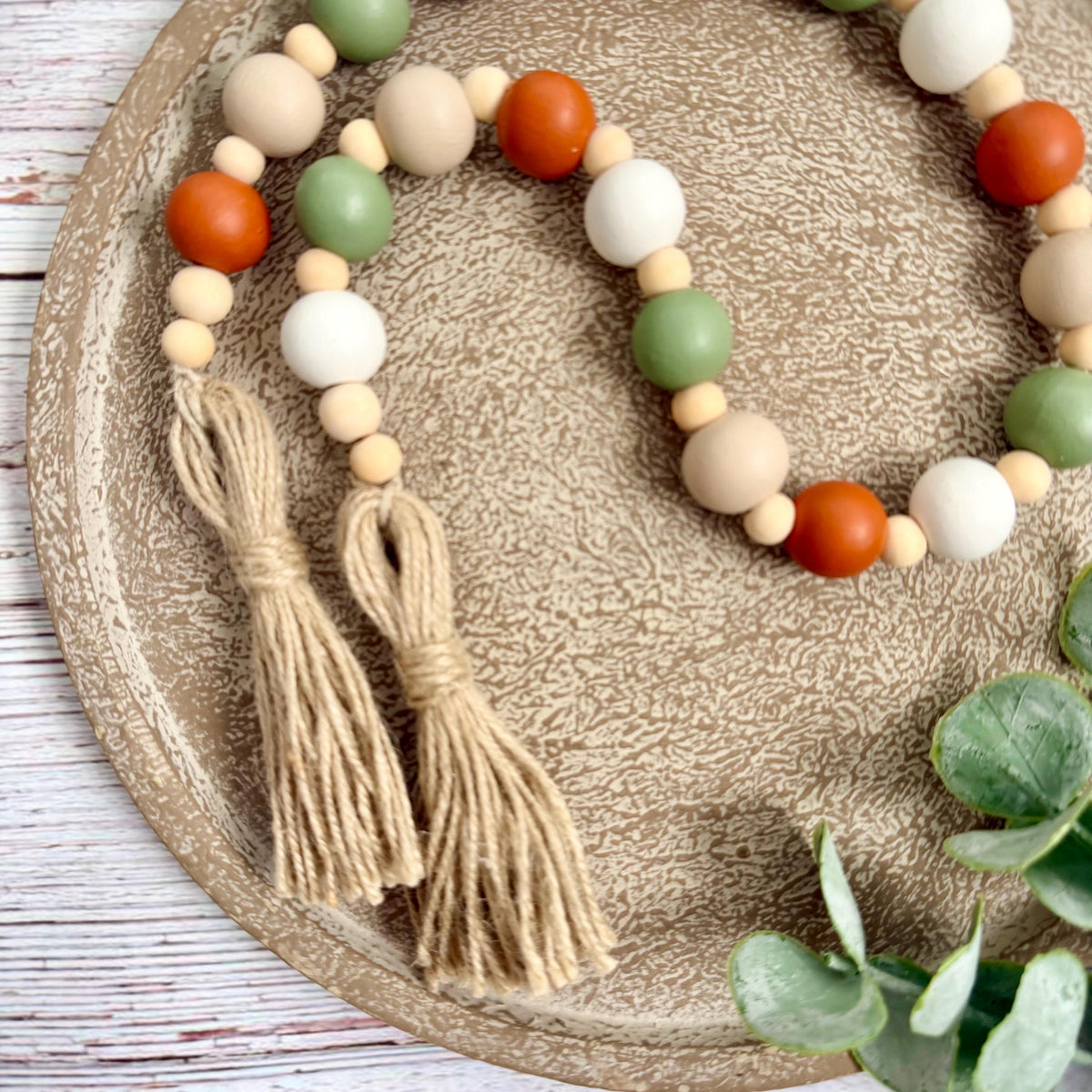 A beaded garland with two tassels, featuring matte wooden beads in terracotta orange, sage green, cream, and white, displayed on a textured beige tray with eucalyptus leaves.