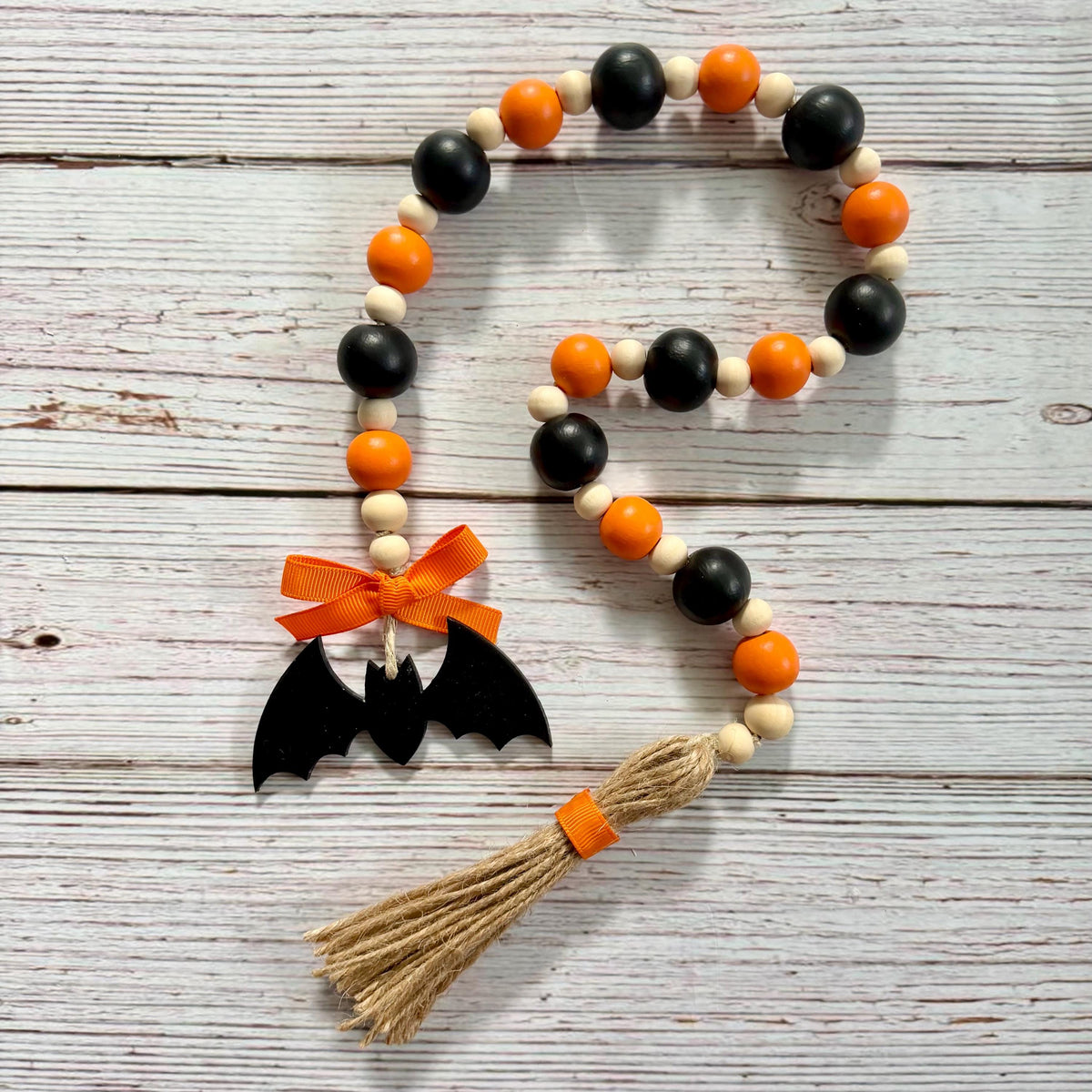 A Halloween-themed wooden bead garland with black and orange beads, a black bat silhouette ornament, an orange ribbon bow, and a jute tassel, displayed on a rustic white-washed wood background.