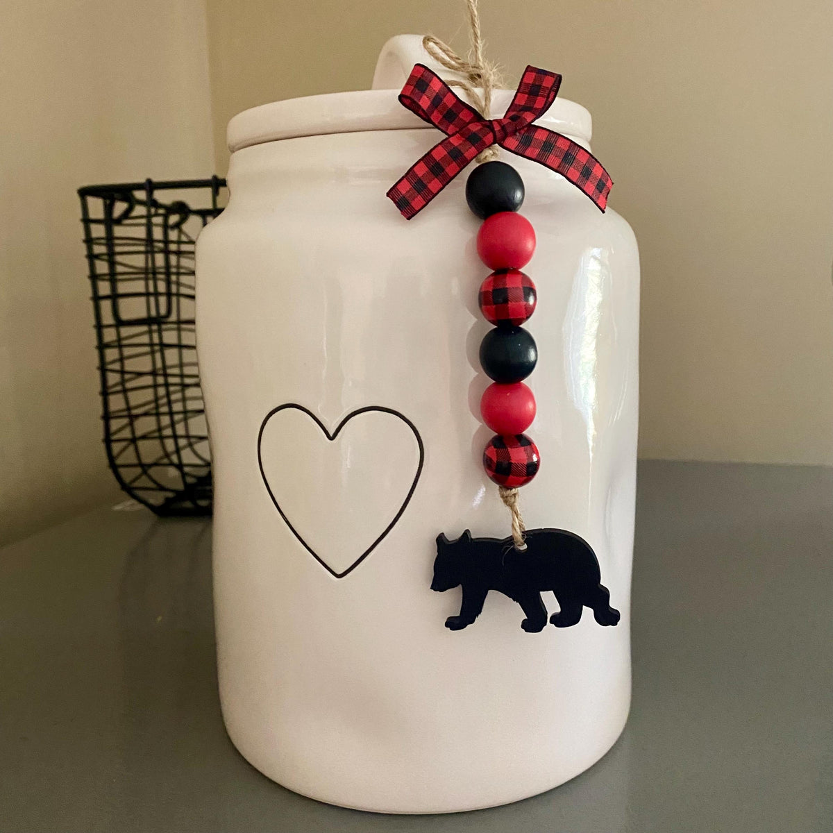 A white ceramic jar with a lid, decorated with a black heart outline, a black bear silhouette, and a hanging beaded garland with red and black plaid accents.