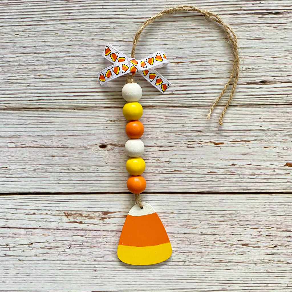 A handmade autumn ornament featuring a wooden candy corn charm at the bottom, a string of white, yellow, and orange beads, a patterned paper bow, and a jute hanging loop, displayed on a rustic white-washed wood background.
