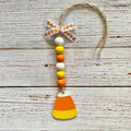 A handmade autumn ornament featuring a wooden candy corn charm at the bottom, a string of white, yellow, and orange beads, a patterned paper bow, and a jute hanging loop, displayed on a rustic white-washed wood background.