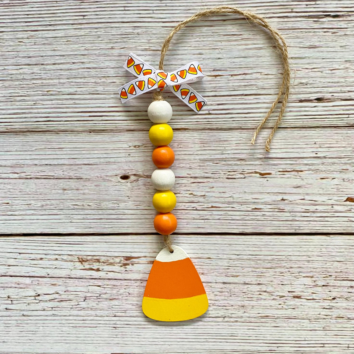 A handmade autumn ornament featuring a wooden candy corn charm at the bottom, a string of white, yellow, and orange beads, a patterned paper bow, and a jute hanging loop, displayed on a rustic white-washed wood background.