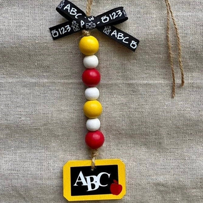 A decorative hanging ornament with a black ribbon printed with white letters and numbers, a vertical string of red, white, and yellow beads, and a yellow tag at the bottom reading ABC with a small red apple.