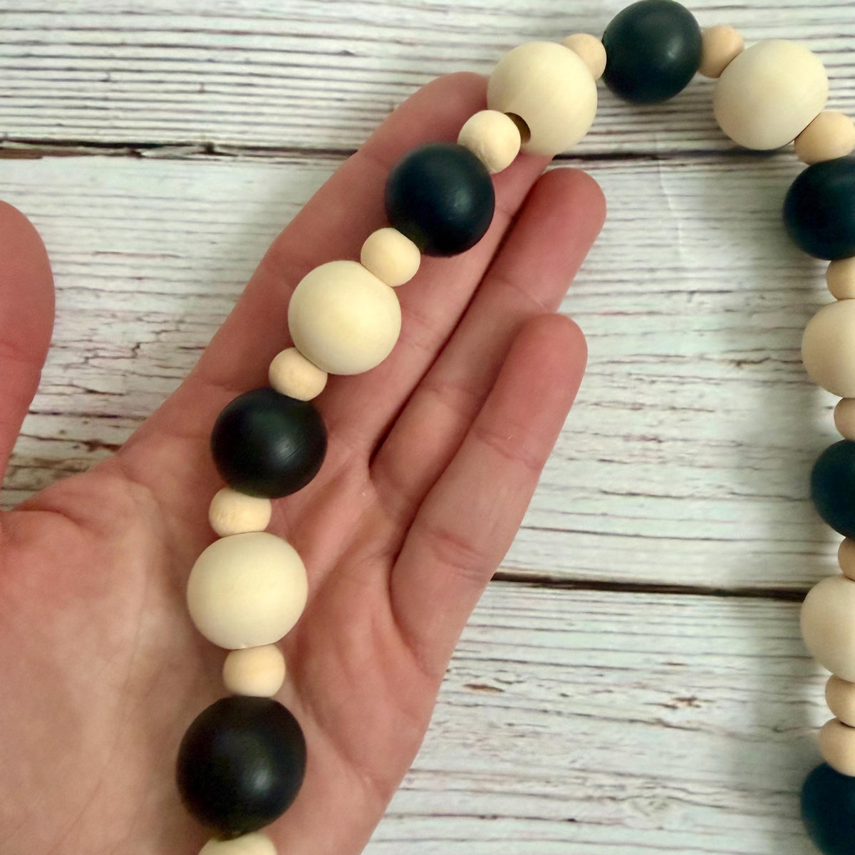 A beaded wooden garland with alternating matte black and cream-colored round beads, held in a person's hand over a whitewashed wooden surface.