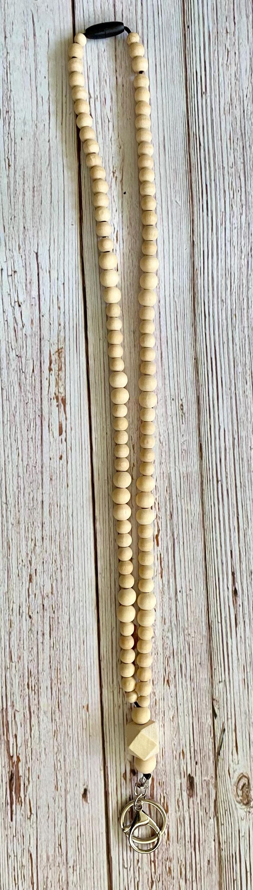 A long strand of light beige wooden beads with a small black connector at the top and a silver-toned metal clasp at the bottom, laid flat on a weathered white wood background.