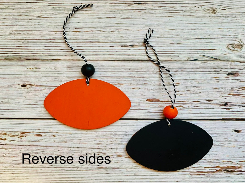 Two oval wall hangings or ornaments displayed on a whitewashed wood background. One is bright orange with a black bead and black-and-white twisted cord, and the other is black with a small orange bead and matching cord.