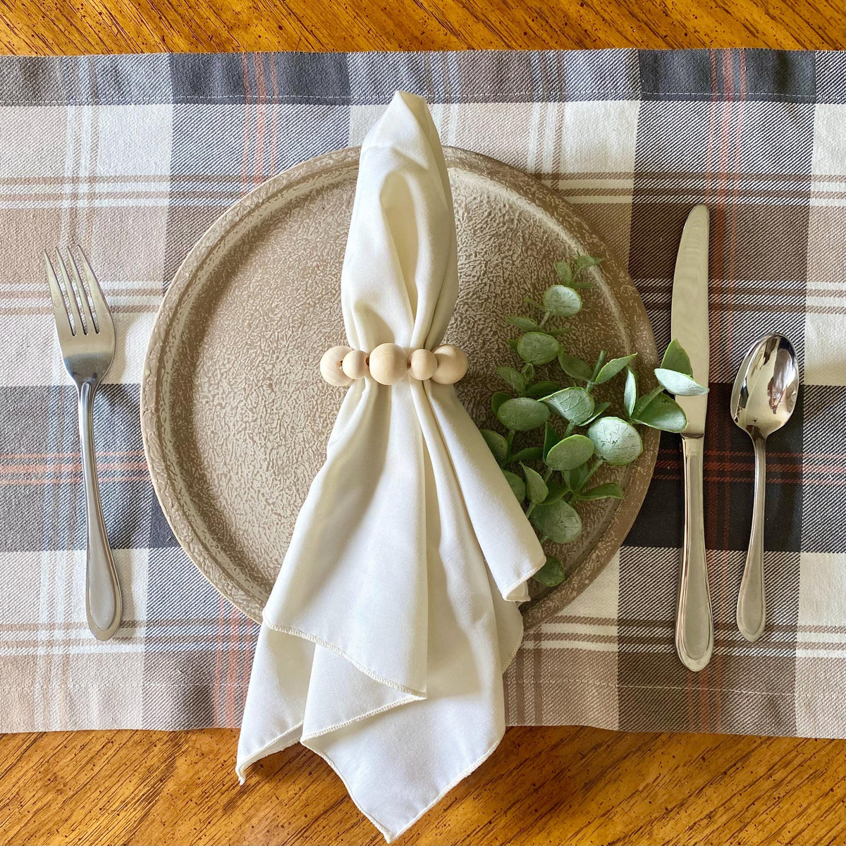 A styled tabletop place setting featuring a beige plaid placemat, a round textured beige plate, a cream cloth napkin tied with wooden beads, green eucalyptus leaves, and silver cutlery on a wooden table.