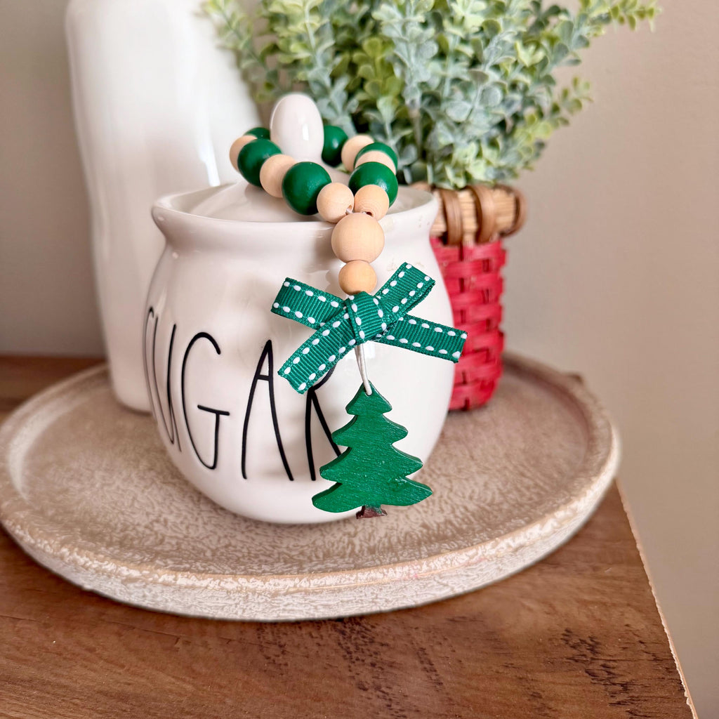 A white ceramic sugar bowl with 'SUGAR' written on it, styled with a green and beige wooden bead garland featuring a green bow and wooden Christmas tree ornament, set on a beige tray on a wooden table.
