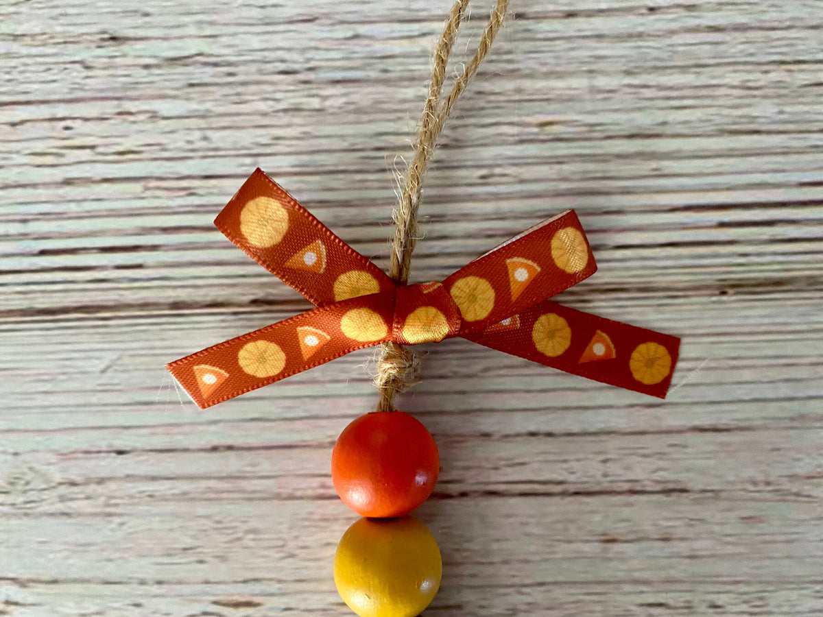 A handmade autumn ornament featuring a red ribbon bow with yellow patterns, a natural jute hanging string, and two wooden beads in red and yellow, displayed on a light gray wood background.