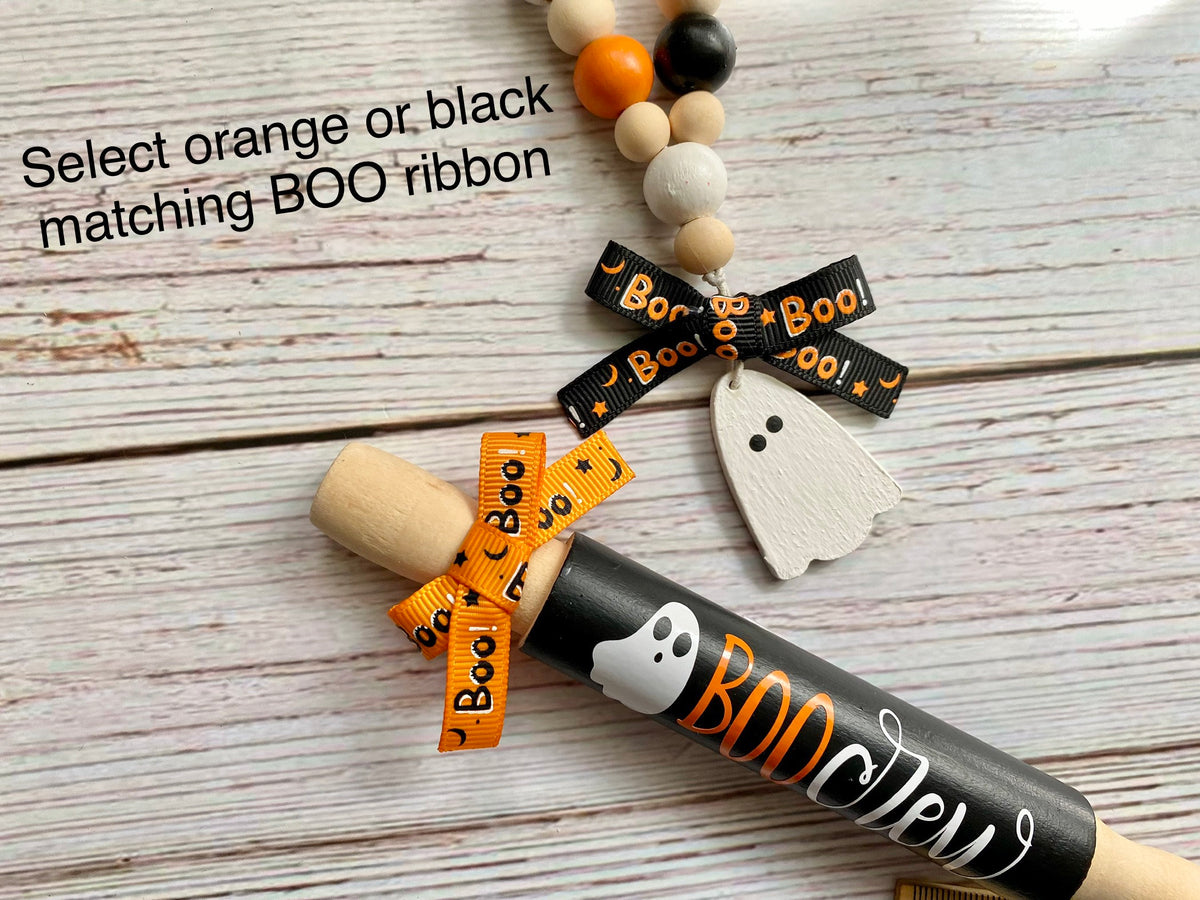 A Halloween-themed wooden rolling pin with a black painted handle, white and orange 'BOO' lettering, and a white ghost charm. It is shown on a whitewashed wood background next to a matching orange ribbon and a black ribbon with 'Boo! Boo! Boo!' text, with text overlay reading 'Select orange or black matching BOO ribbon'.