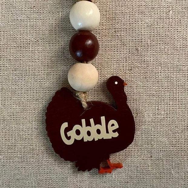 A rustic wooden turkey ornament with the word 'Gobble' cut out from the front, hanging from a beaded string with cream and dark brown beads, set against a textured beige linen background.