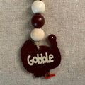 A rustic wooden turkey ornament with the word 'Gobble' cut out from the front, hanging from a beaded string with cream and dark brown beads, set against a textured beige linen background.