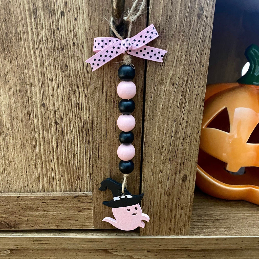 A handmade Halloween hanging ornament with pink and black wooden beads, a pink polka-dot bow, and a ghost charm, displayed on a rustic wooden shelf next to a jack-o'-lantern.