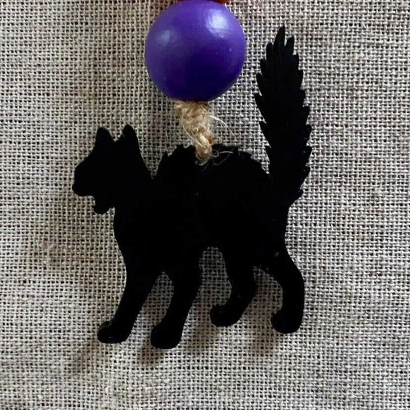 A black cat silhouette ornament with a purple spherical bead and natural jute twine, displayed on a textured beige linen background.