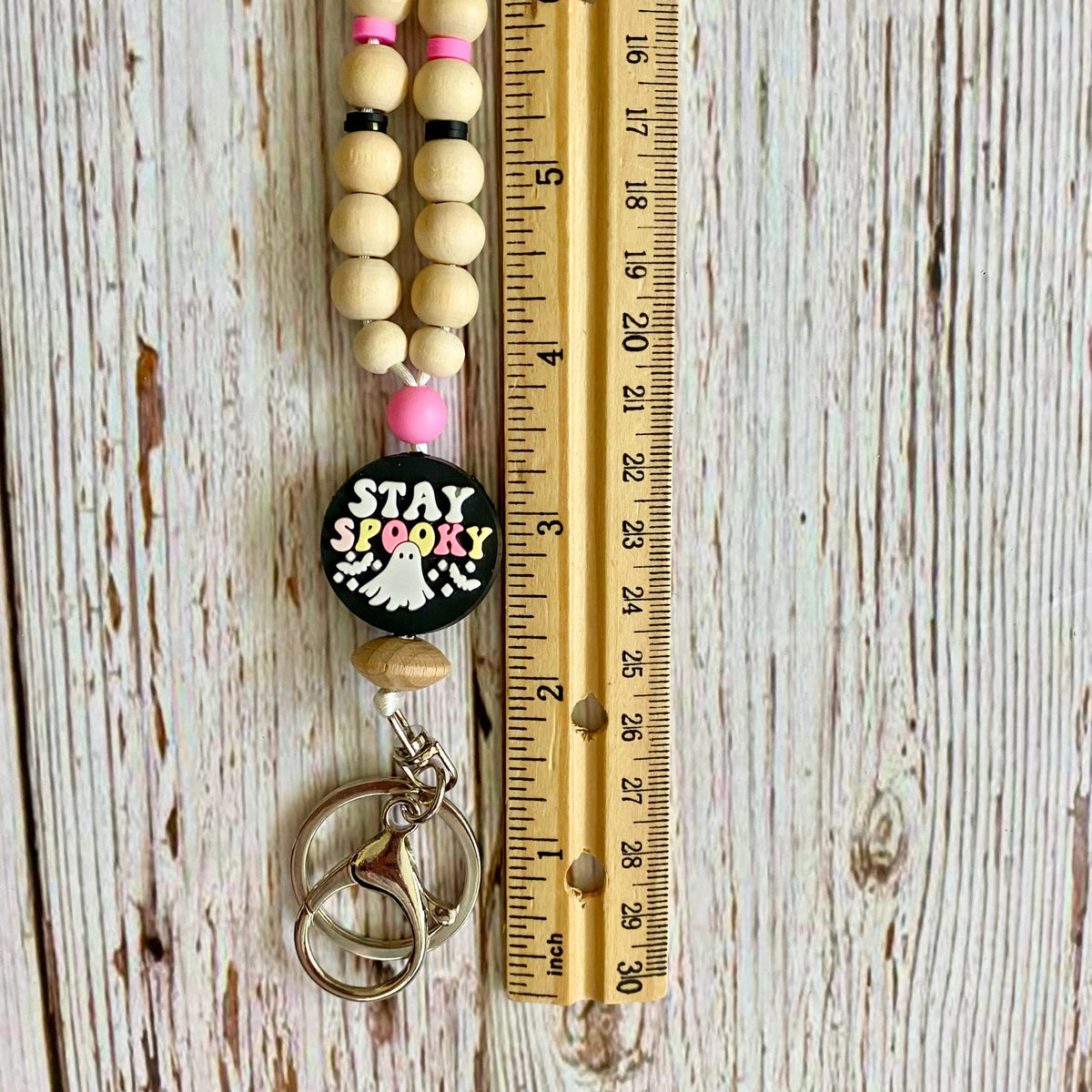 A beaded keychain with a black round charm that says 'STAY SPOOKY' in colorful letters, shown next to a wooden ruler on a whitewashed wood background.