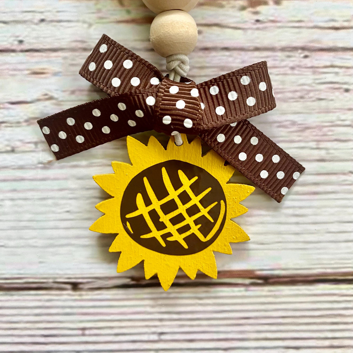 A decorative sunflower-shaped ornament with a yellow face, dark brown center, and a brown ribbon bow with white polka dots tied above it, hanging against a whitewashed wood plank background.