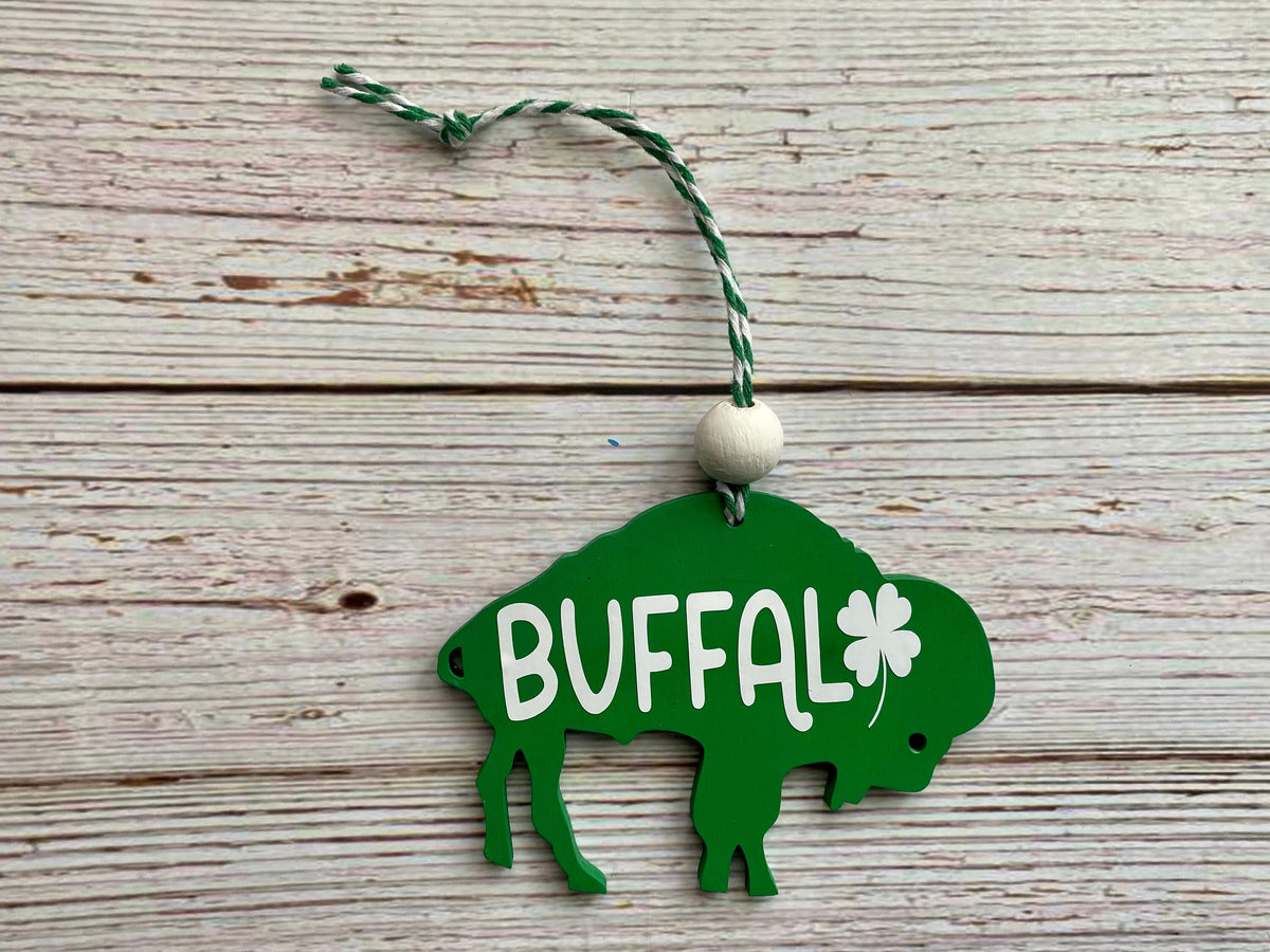 A green buffalo-shaped hanging ornament with the word BUFFAL in white and a white shamrock, suspended by a green-and-white string on a light wood plank background.