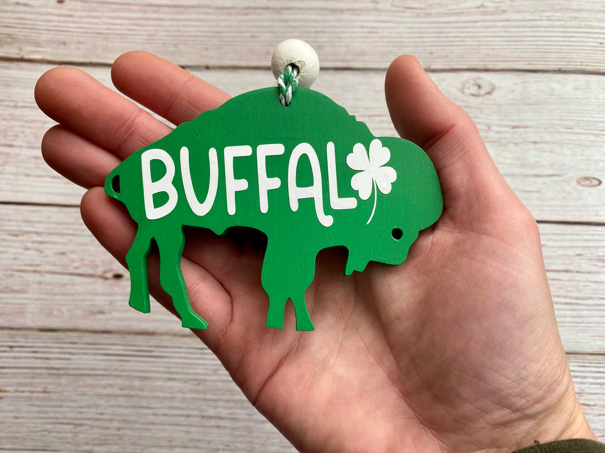 A hand holding a green buffalo-shaped ornament with the word 'BUFFALO' in white letters and a white shamrock, against a light wood plank background.