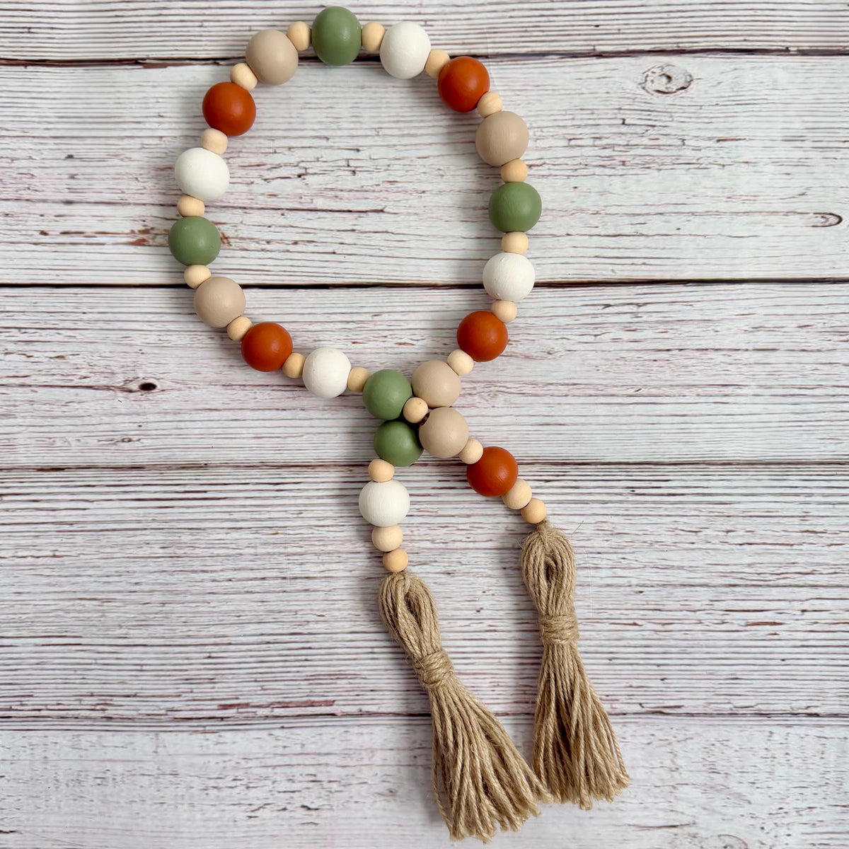 A beaded garland with matte wooden beads in sage green, white, tan, and terracotta, featuring two long jute tassels, displayed on a whitewashed wooden background.