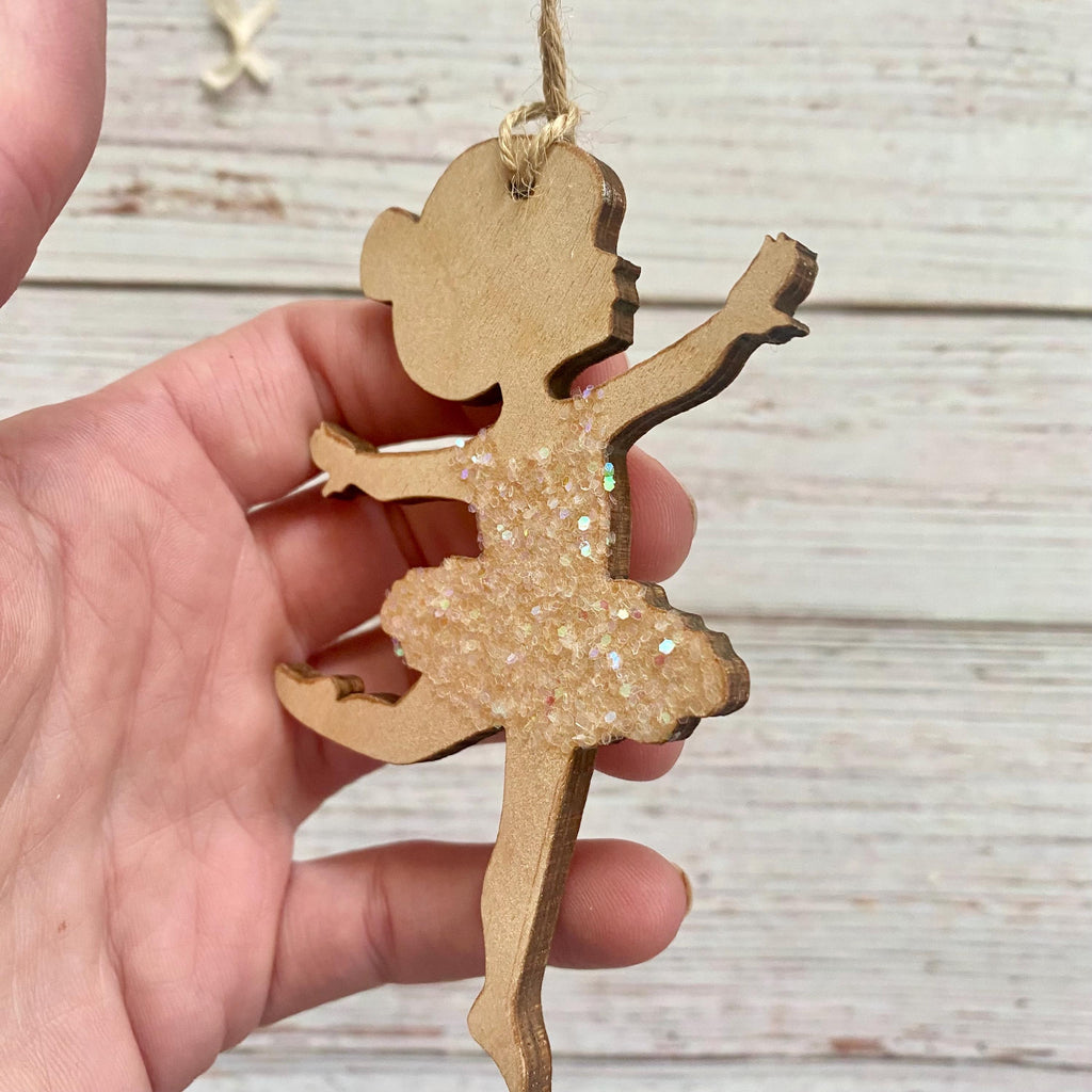 A hand holds a wooden ballerina ornament with a glittery beige dress and a natural wood head and limbs, suspended by a string against a whitewashed wood background.