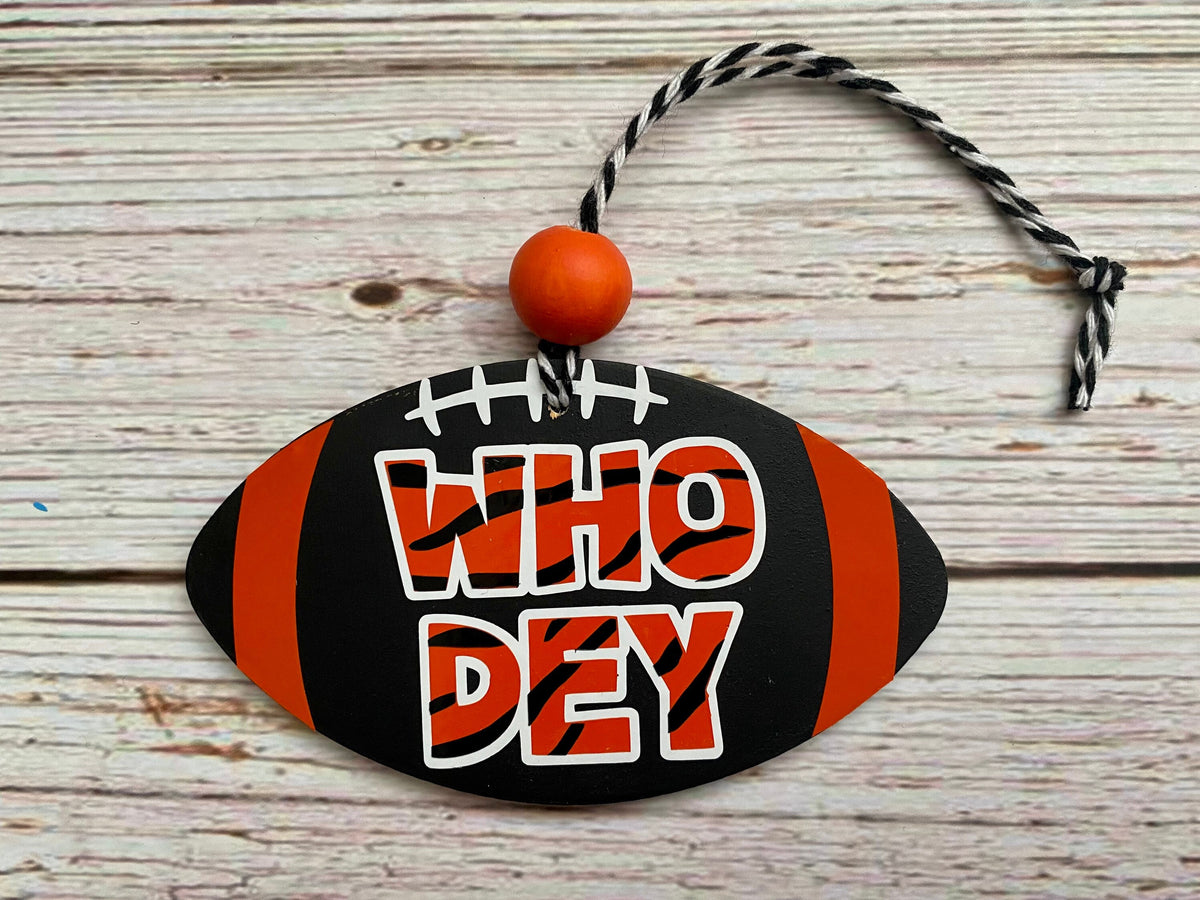 A football-shaped wooden ornament with orange and black stripes, white laces, and the text 'WHO DEY' in orange and white, hanging from a black-and-white twisted string with an orange bead, set on a light wood plank background.