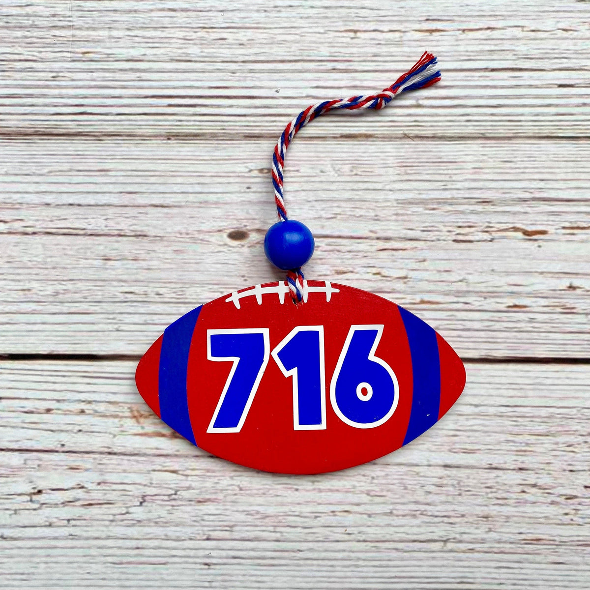 A football-shaped wooden ornament with the numbers 716 in blue and white, featuring a red body, blue side stripes, white faux laces, and a blue hanging bead with a red, white, and blue tassel, displayed on a rustic white wood background.