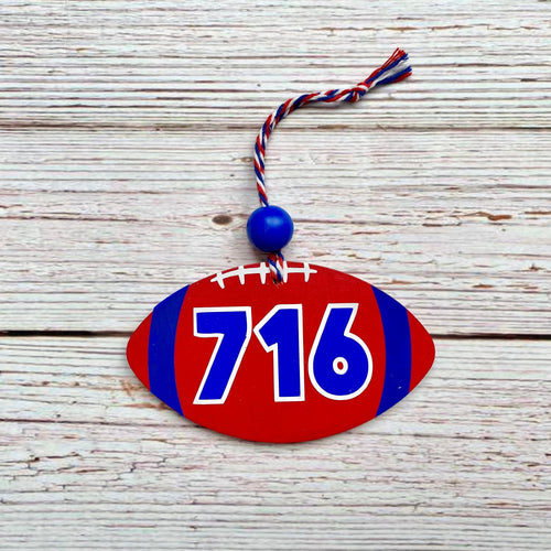 A football-shaped wooden ornament with the numbers 716 in blue and white, featuring a red body, blue side stripes, white faux laces, and a blue hanging bead with a red, white, and blue tassel, displayed on a rustic white wood background.