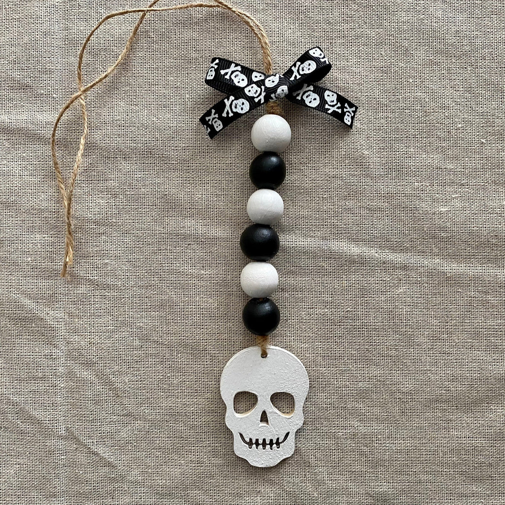 A Halloween ornament featuring a white skull charm, black and white wooden beads, a black and white skull-patterned ribbon bow, and a jute hanger, displayed on a textured beige linen background.