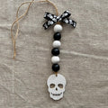 A Halloween ornament featuring a white skull charm, black and white wooden beads, a black and white skull-patterned ribbon bow, and a jute hanger, displayed on a textured beige linen background.