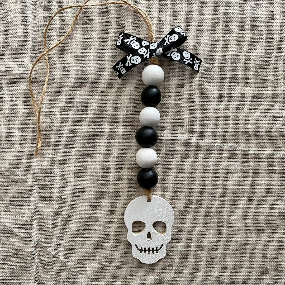A Halloween ornament featuring a white skull charm, black and white wooden beads, a black and white skull-patterned ribbon bow, and a jute hanger, displayed on a textured beige linen background.