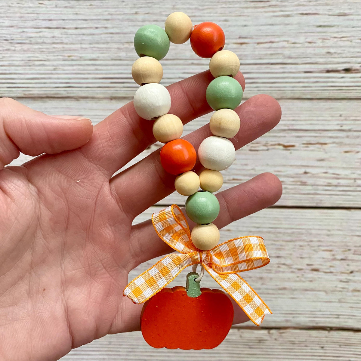 A hand holds a beaded loop with orange, mint green, cream, and white wooden beads, finished with an orange pumpkin charm and an orange-and-white gingham ribbon bow, against a whitewashed wood background.
