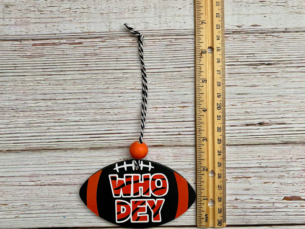 A football-shaped hanging ornament with the phrase 'WHO Dey' in red and white lettering, black and orange stripes, and a black-and-white string, shown next to a wooden ruler on a whitewashed wood background.